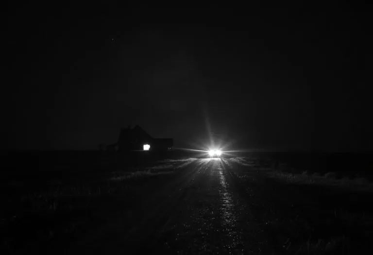 Two men sit in the front seat of a car at night, their faces lit by dashboard light and shadows, rain streaking the