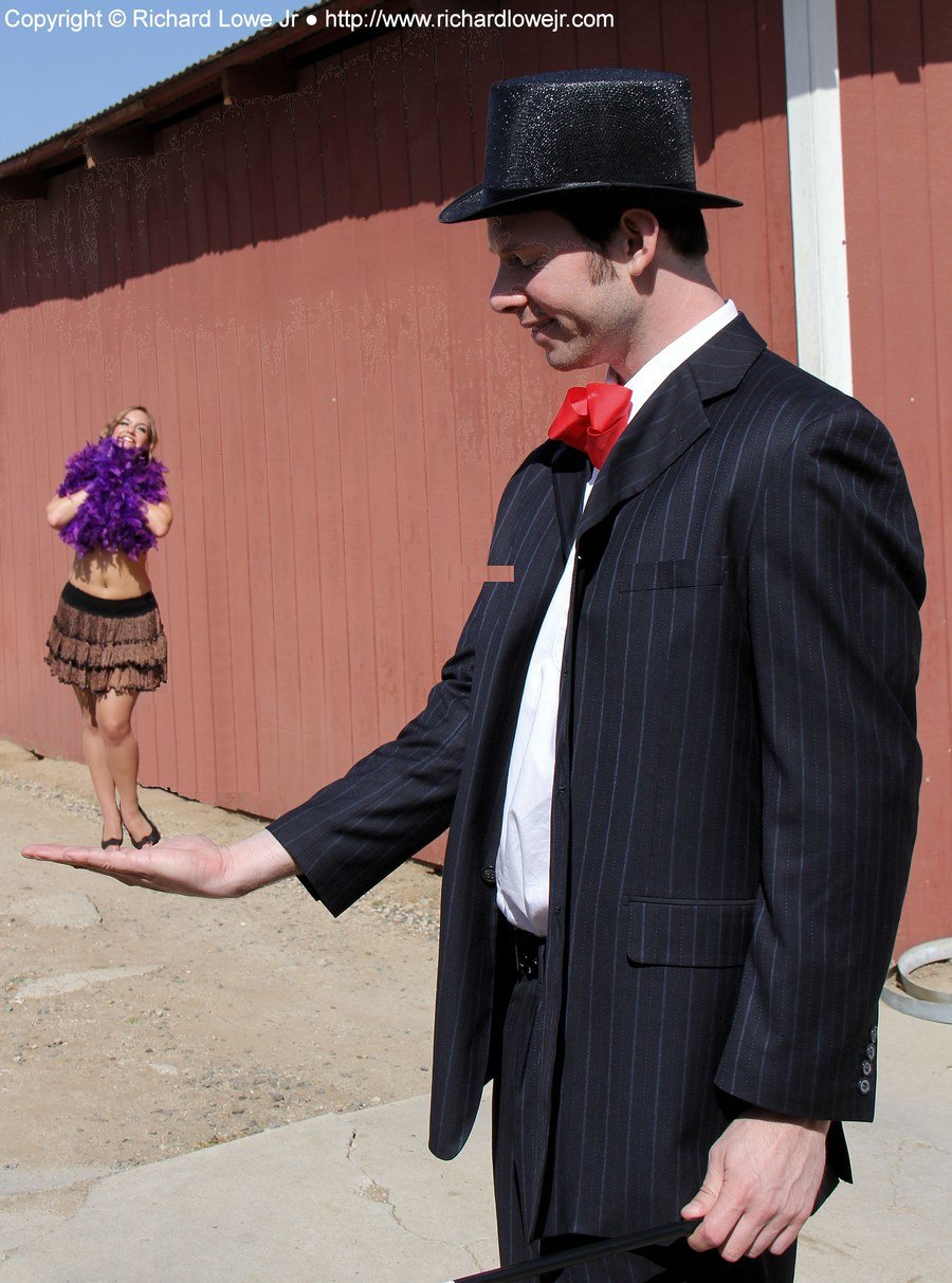 Forced perspective photo of a ringmaster in a top hat and pinstripe suit who appears to hold a tiny dancer with a purple feather boa in the palm of his hand