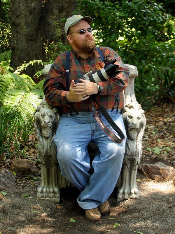Gar Travis in a plaid shirt, cap and sunglasses sitting on a stone bench carved with lions, holding a camera with a long lens