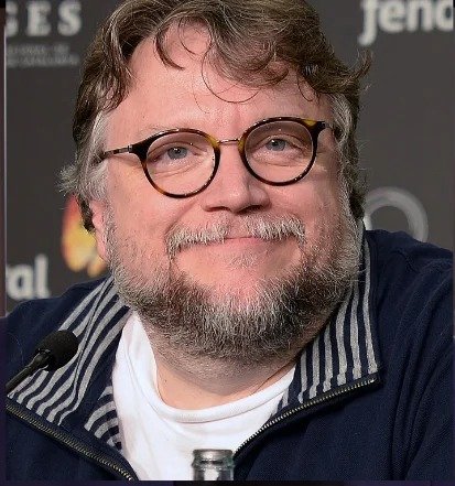 Guillermo del Toro smiling at a press conference in 2017, wearing round glasses and a dark zip jacket