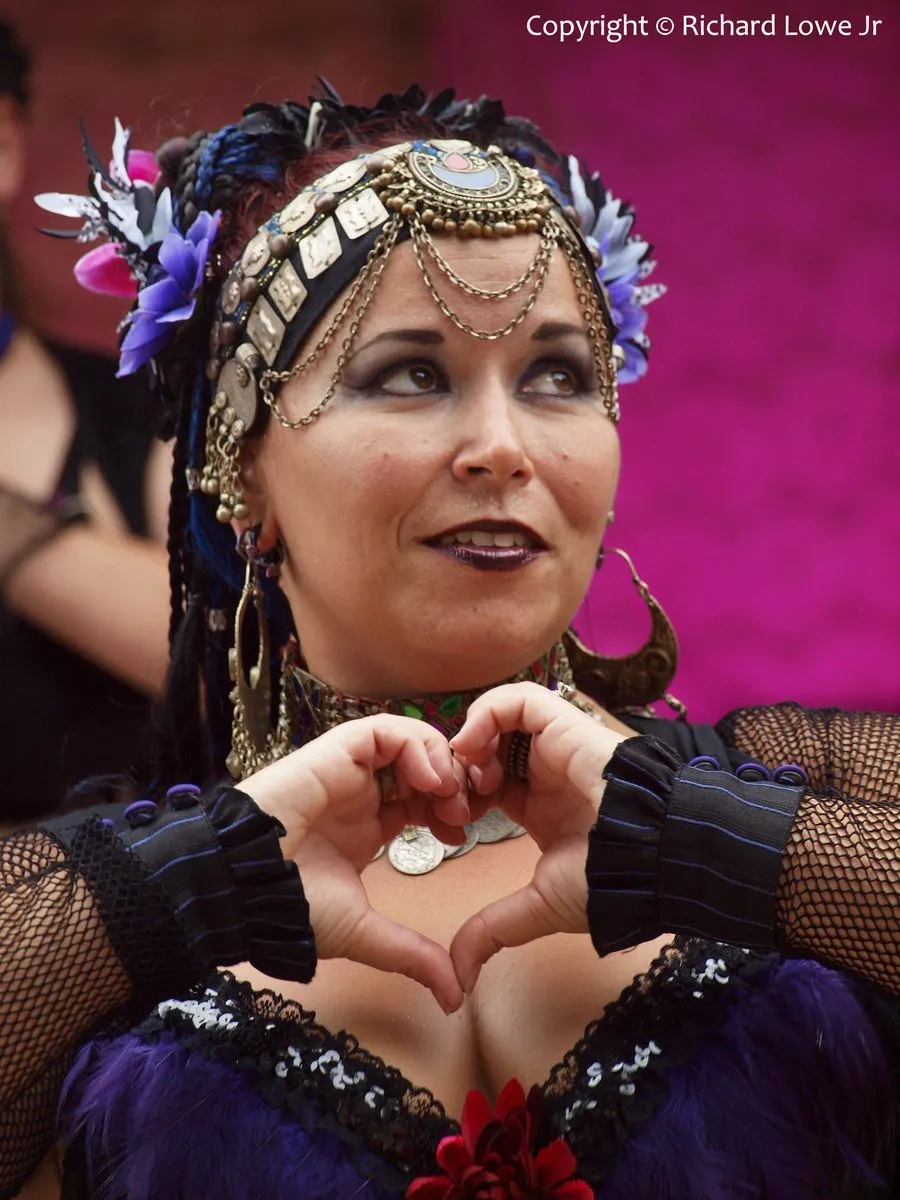 Tribal belly dancer Karma in a coin headdress and chain jewelry, glancing up with a smile and making a heart with her hands, at Ojai Day