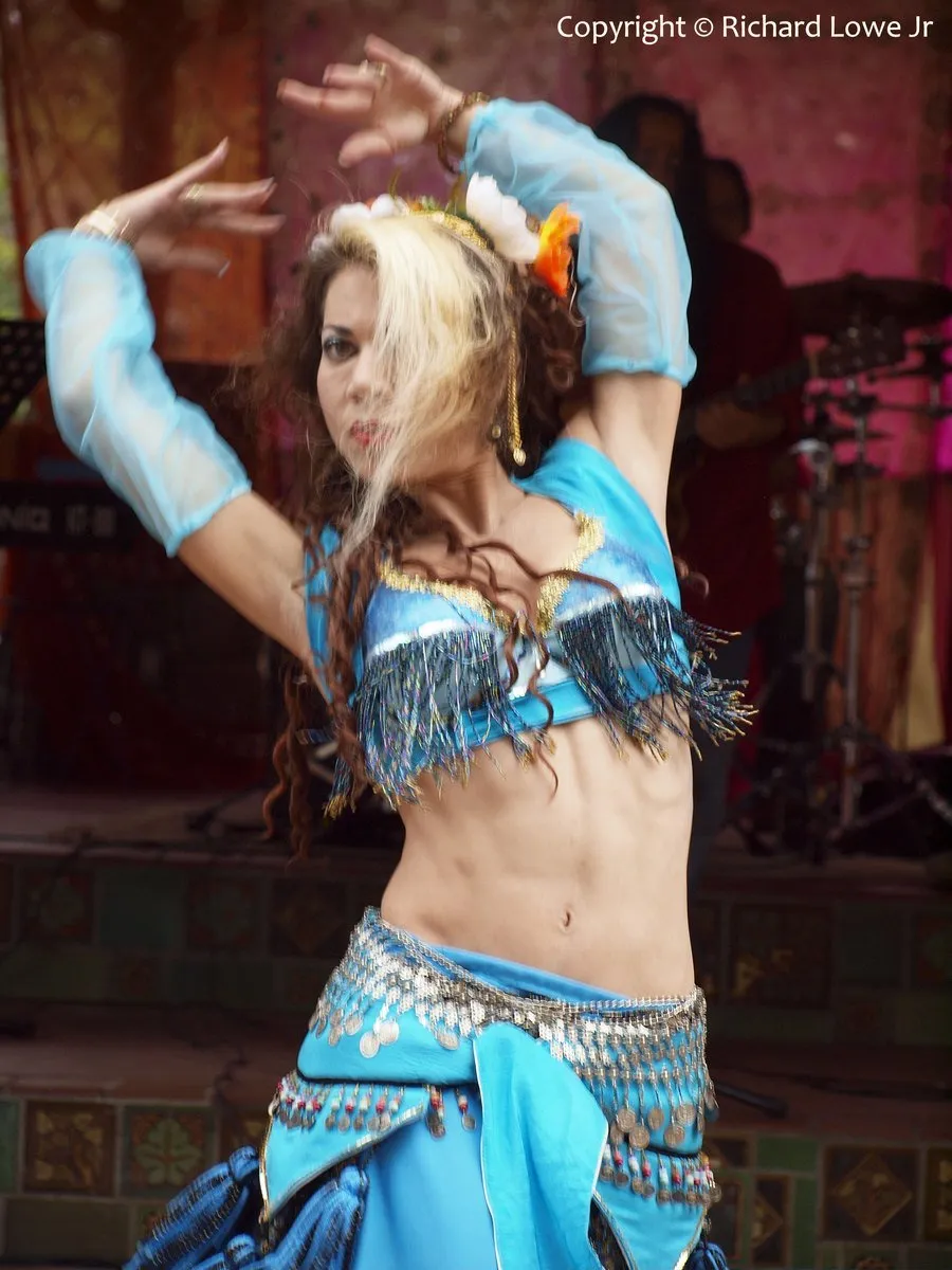 Belly dancer Nilay in a turquoise beaded costume, arms raised and hair flying mid-turn, dancing in front of a live band at Ojai Day