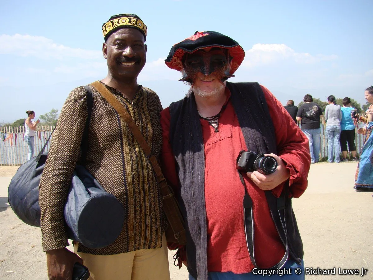 Vernon Lewis in an embroidered cap and patterned tunic carrying a drum bag, smiling beside Richard Lowe in a red shirt, leather mask and faire hat holding a camera at the Irwindale Renaissance Faire