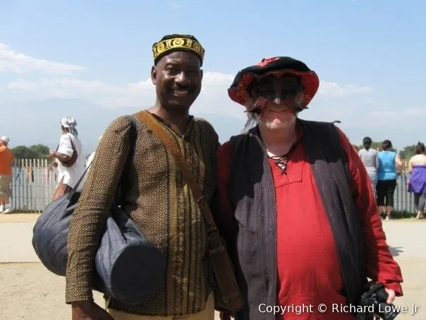 Vernon Lewis in an embroidered cap and tunic standing beside Richard Lowe in faire costume and a feathered hat at the Irwindale Renaissance Faire